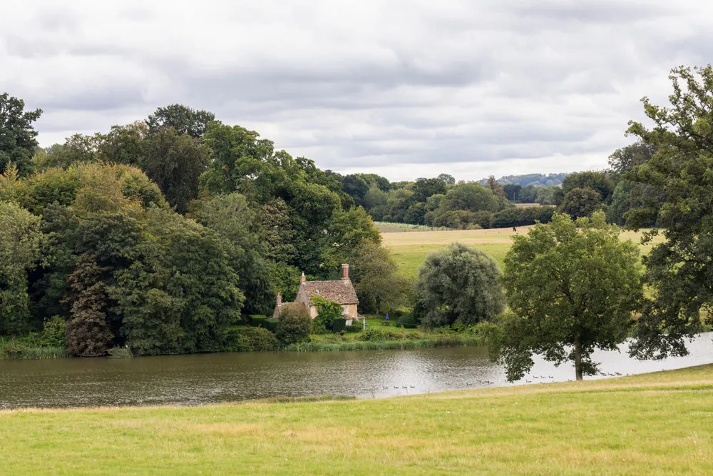 A small cottage surrounded by dense trees is situated near the edge of a large pond in a rural landscape on a cloudy day.