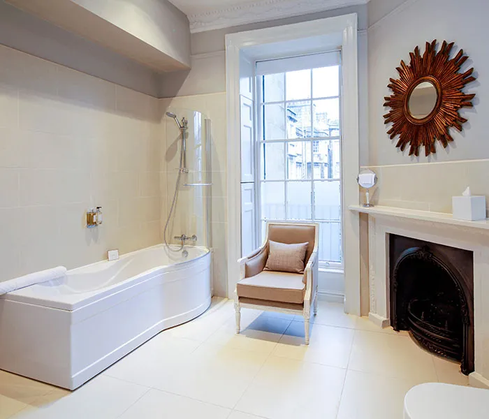 A spacious bathroom with a bathtub, a small armchair, a fireplace, a sunburst mirror, and a large window with natural light.