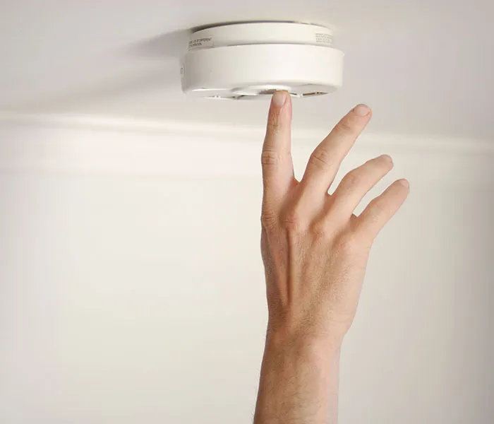 A hand is reaching up to a ceiling-mounted smoke detector.