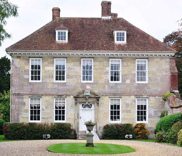 A symmetrical, two-story stone house with a red-tiled roof and a white front door, surrounded by a manicured lawn and bushes.