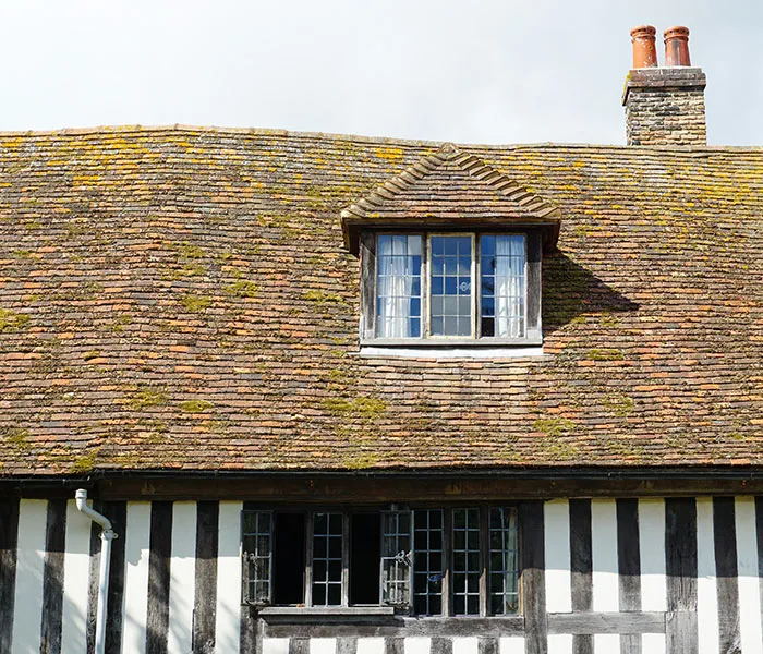 A traditional half-timbered house with a tiled roof, featuring a dormer window and a brick chimney, is shown under a clear sky.