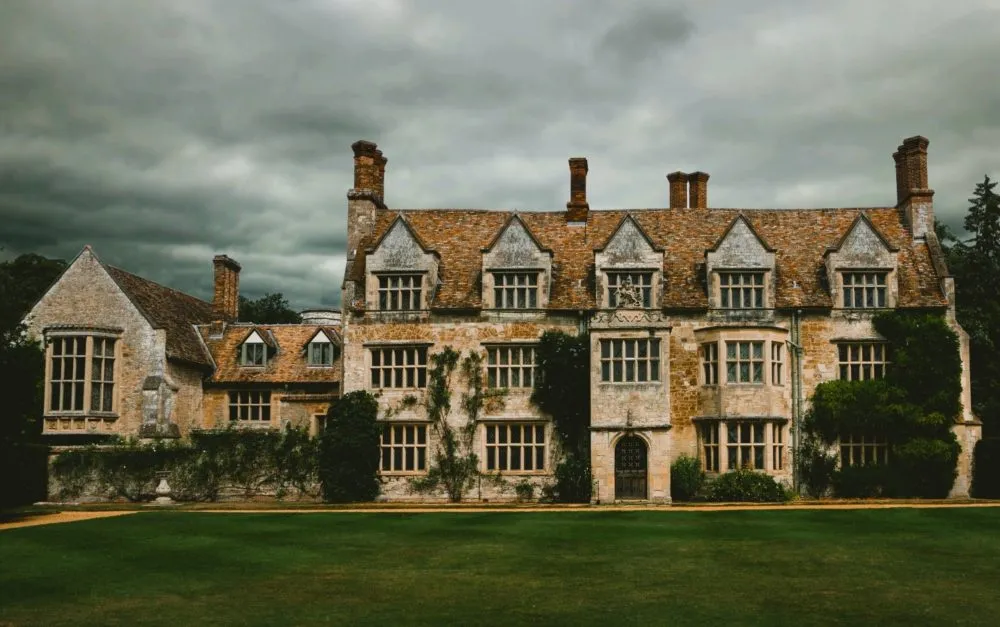 A large, old stone manor with ivy-covered walls, numerous chimneys, and a cloudy sky overhead.