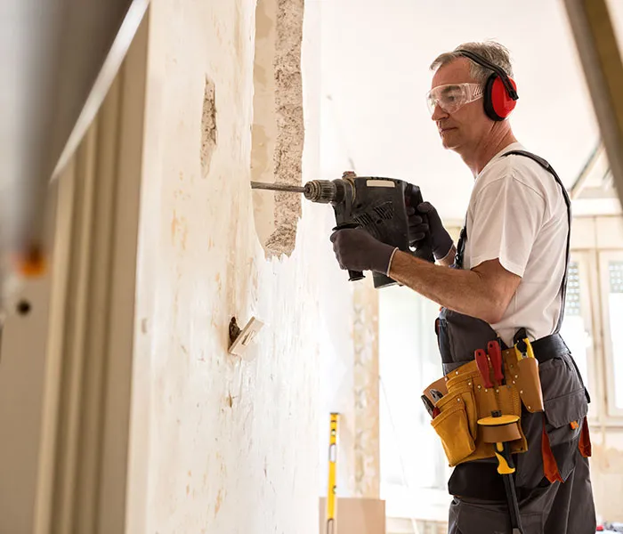 A man wearing safety gear uses a power drill on a wall inside a partially renovated room. He has tools attached to his belt.