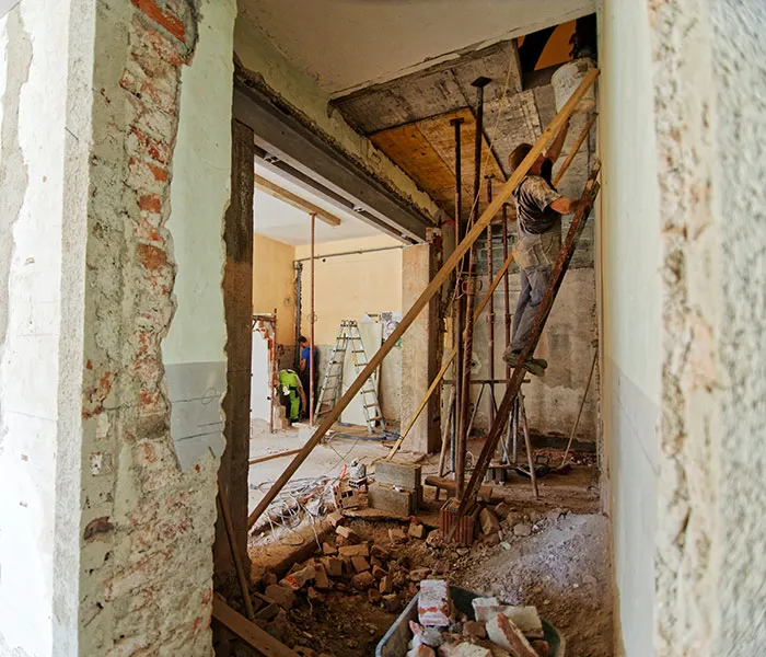 Construction worker on scaffolding in a partially demolished building under renovation, with tools and debris scattered on the floor.