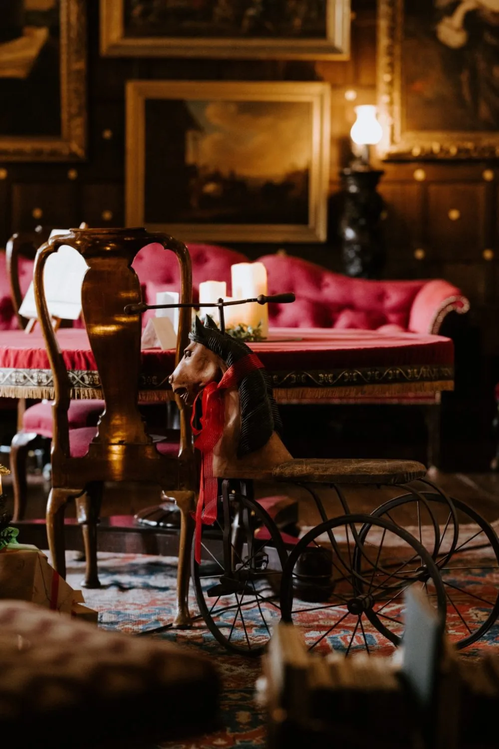 A vintage toy horse on a tricycle sits in front of a wooden chair, with a red couch and framed artwork in the background.