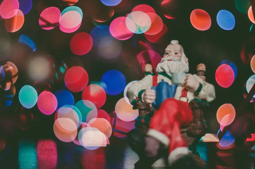 A figurine of Santa Claus sitting on a rocking chair, with colorful blurred lights in the background.