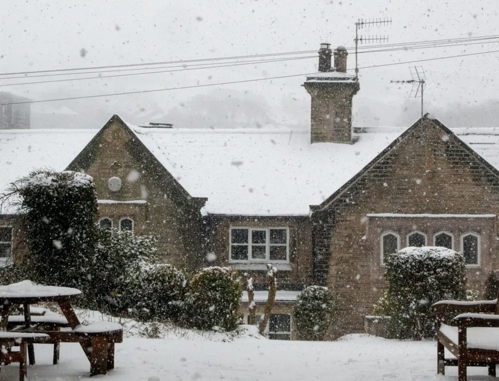 Heavy snow falls on a row of old stone houses and surrounding trees with snow-covered picnic tables in the foreground.