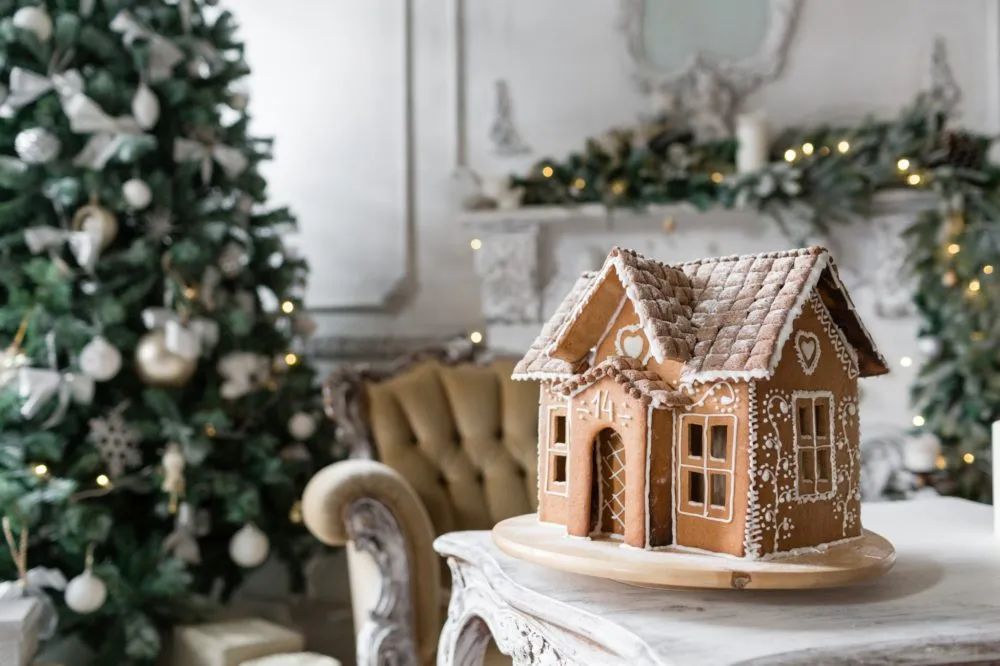 A gingerbread house sits on a white table with a decorated Christmas tree and vintage armchair in the background.