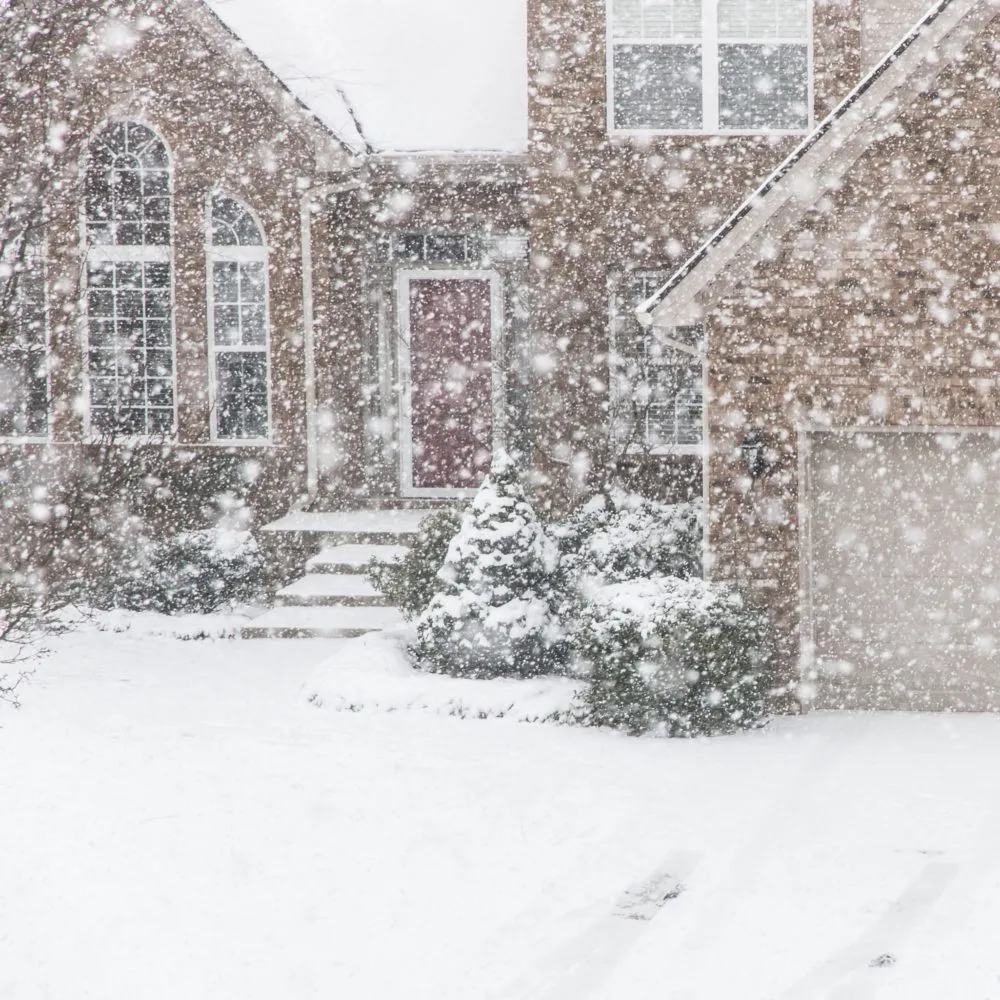 A brick house with snow-covered trees and bushes in front, as heavy snowfall blankets the scene, partially obscuring visibility.