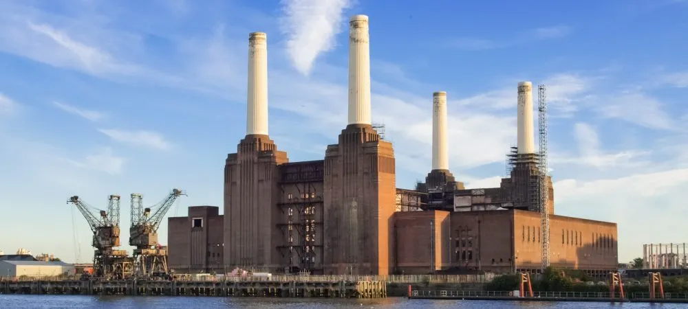 A large brick power station with four tall white chimneys, surrounded by cranes and scaffolding, under a blue sky with light clouds.