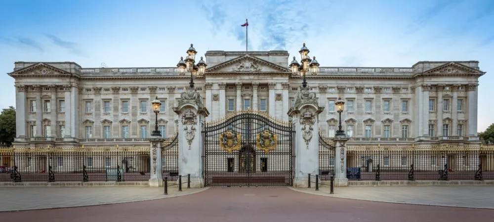 Front view of Buckingham Palace with its ornate gates, adorned lampposts, and a flag flying at the top, under a clear sky.