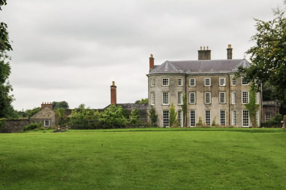 A large, three-story historic stone mansion with tall windows stands on a well-manicured lawn under an overcast sky.