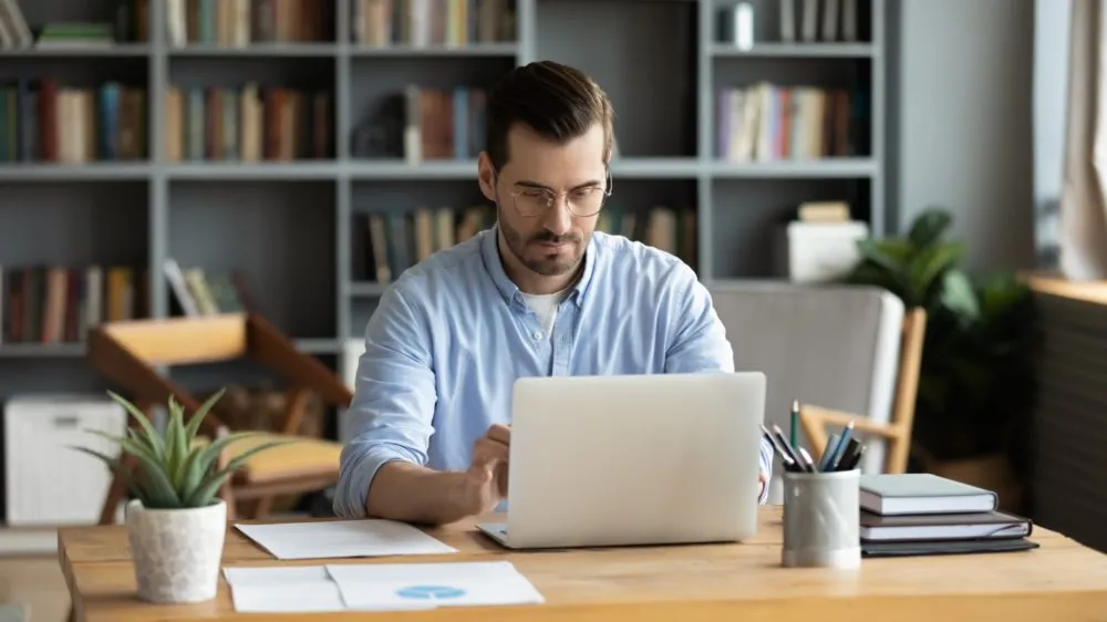 A man wearing glasses works on a laptop at a wooden desk in a home office with bookshelves in the background.