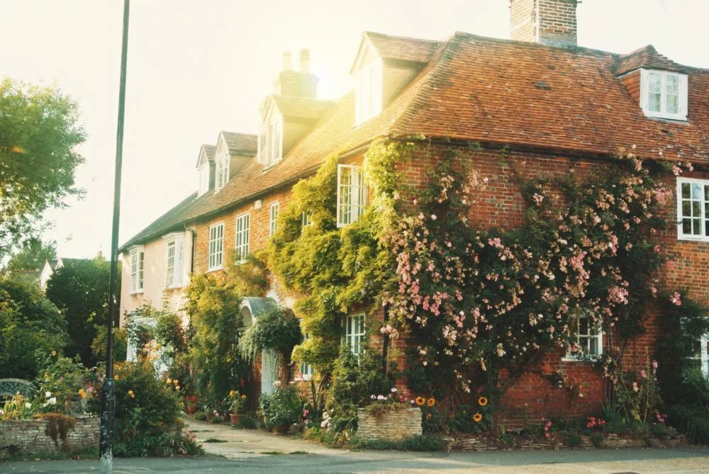 A traditional brick house with dormer windows and a red tile roof, covered in climbing plants and surrounded by a lush garden.