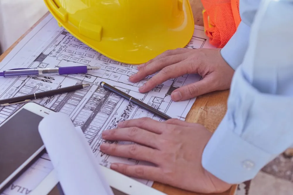 Person's hands on architectural blueprints with a yellow hard hat, pens, and tablet on the table.