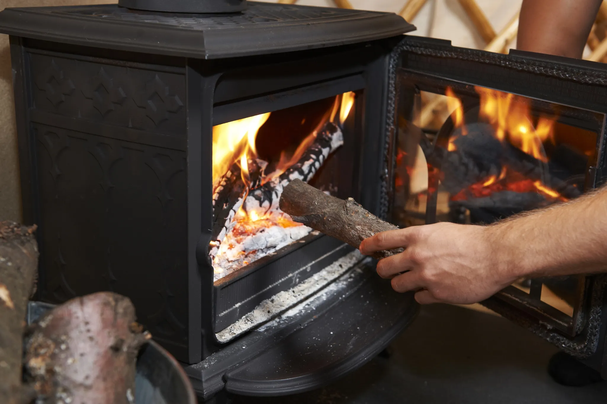 A person placing a log into a burning wood stove, with flames visible inside and the stove door open.