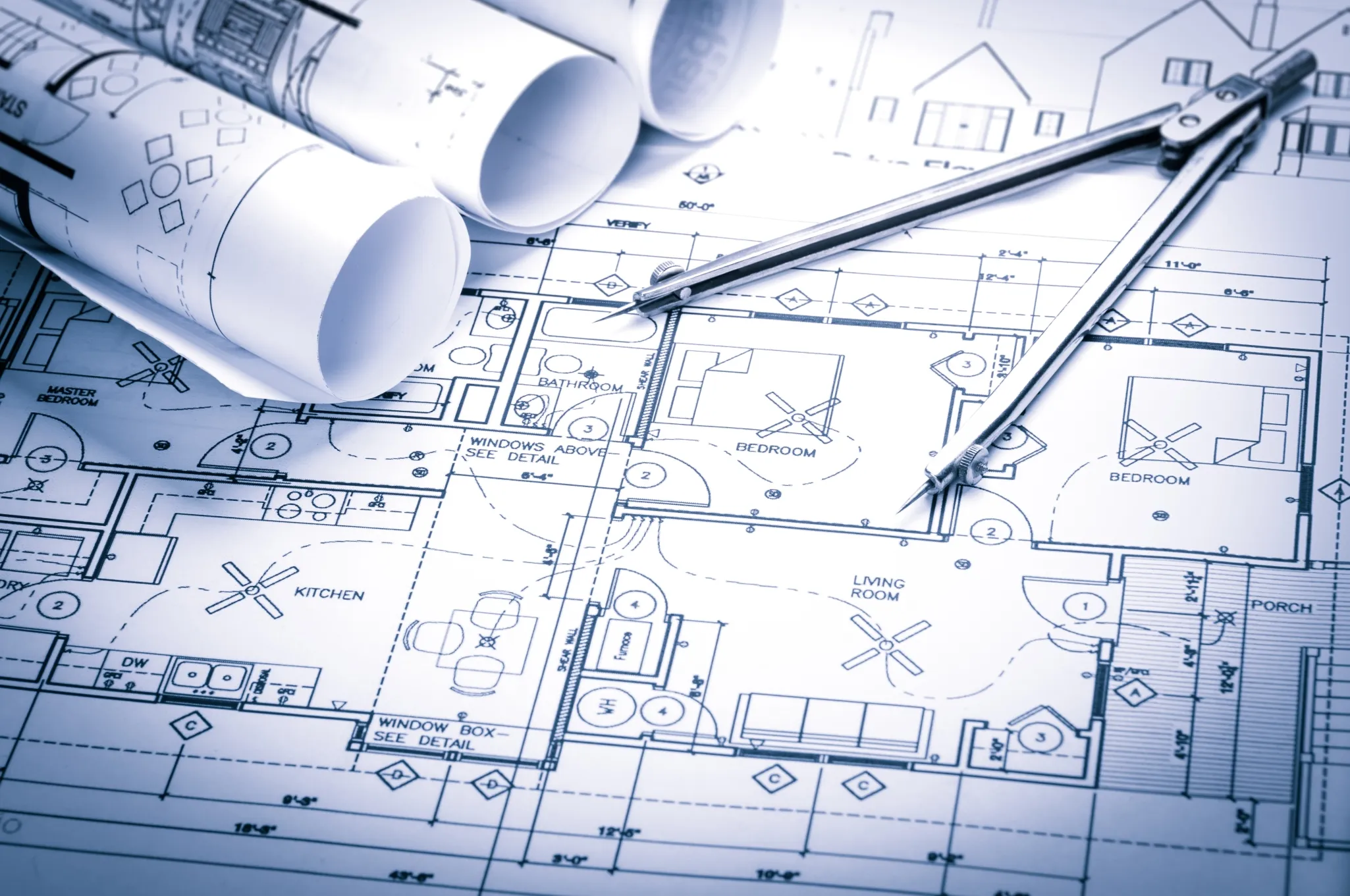 Blueprints of a house layout with architectural drawings, rolled-up plans, and a pair of compasses on top.