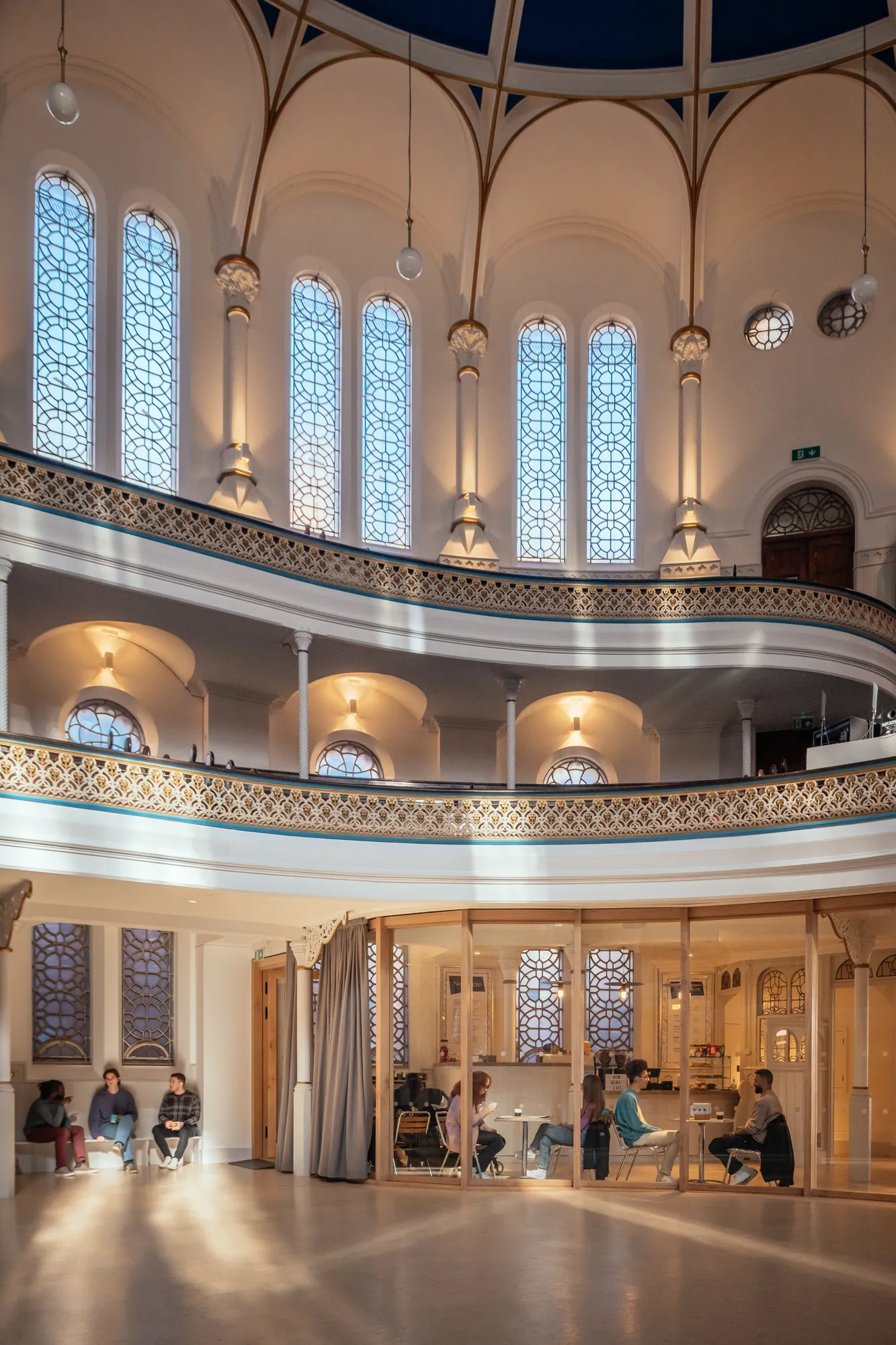 A spacious, well-lit interior with high arched windows and people seated in groups on the lower level of the two-tiered structure.