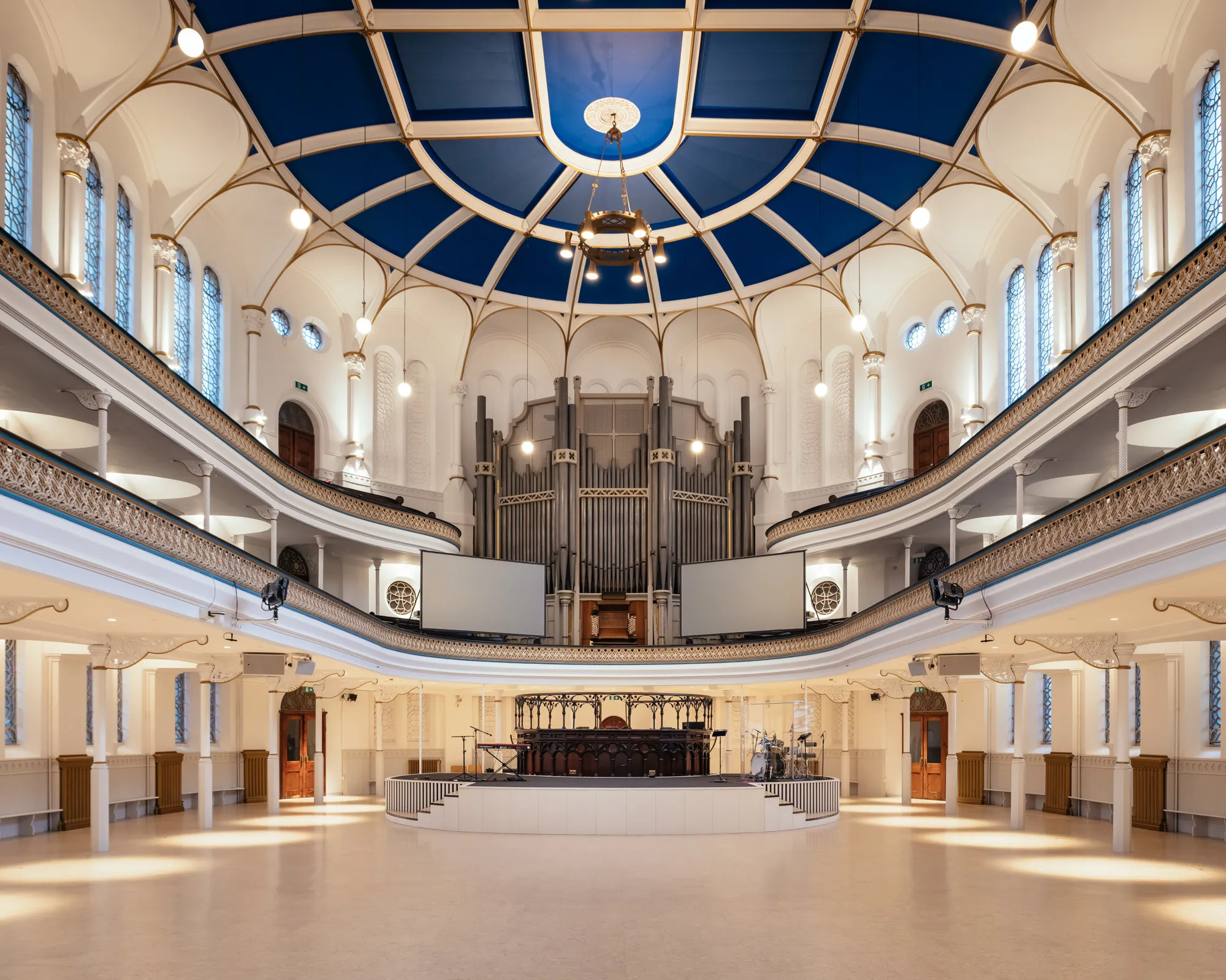 A wide view of an ornate concert hall interior with a large pipe organ, a blue and white vaulted ceiling, and balcony seating.