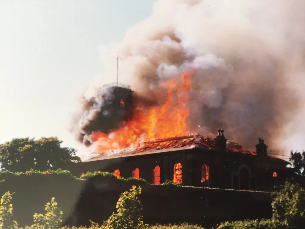 A large building engulfed in flames with thick smoke billowing into the sky, surrounded by greenery in daylight.