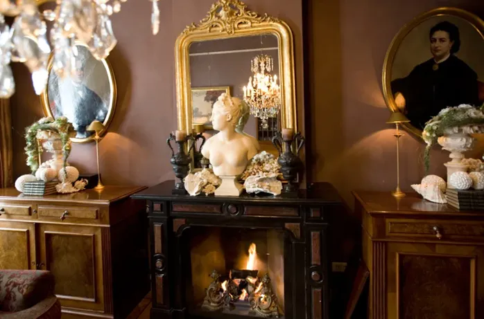 An ornate fireplace with a lit fire, topped by a bust statue, flanked by framed portraits and decorated with seashell displays.