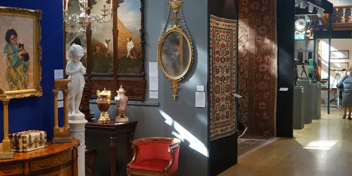 A gallery room featuring antique furniture, a statue, paintings, and ornate rugs displayed in an exhibition setting.