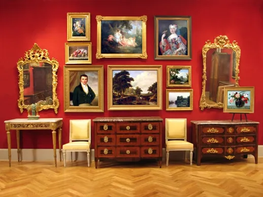 A gallery room with red walls, showcasing ornate paintings and mirrors above antique wooden furniture with marble tops and yellow chairs.