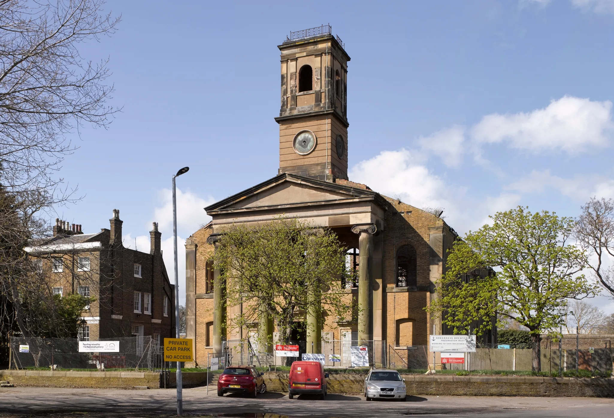 An old, partially restored church building with a clock tower, surrounded by fencing, with parked cars in front. Multiple signs visible.