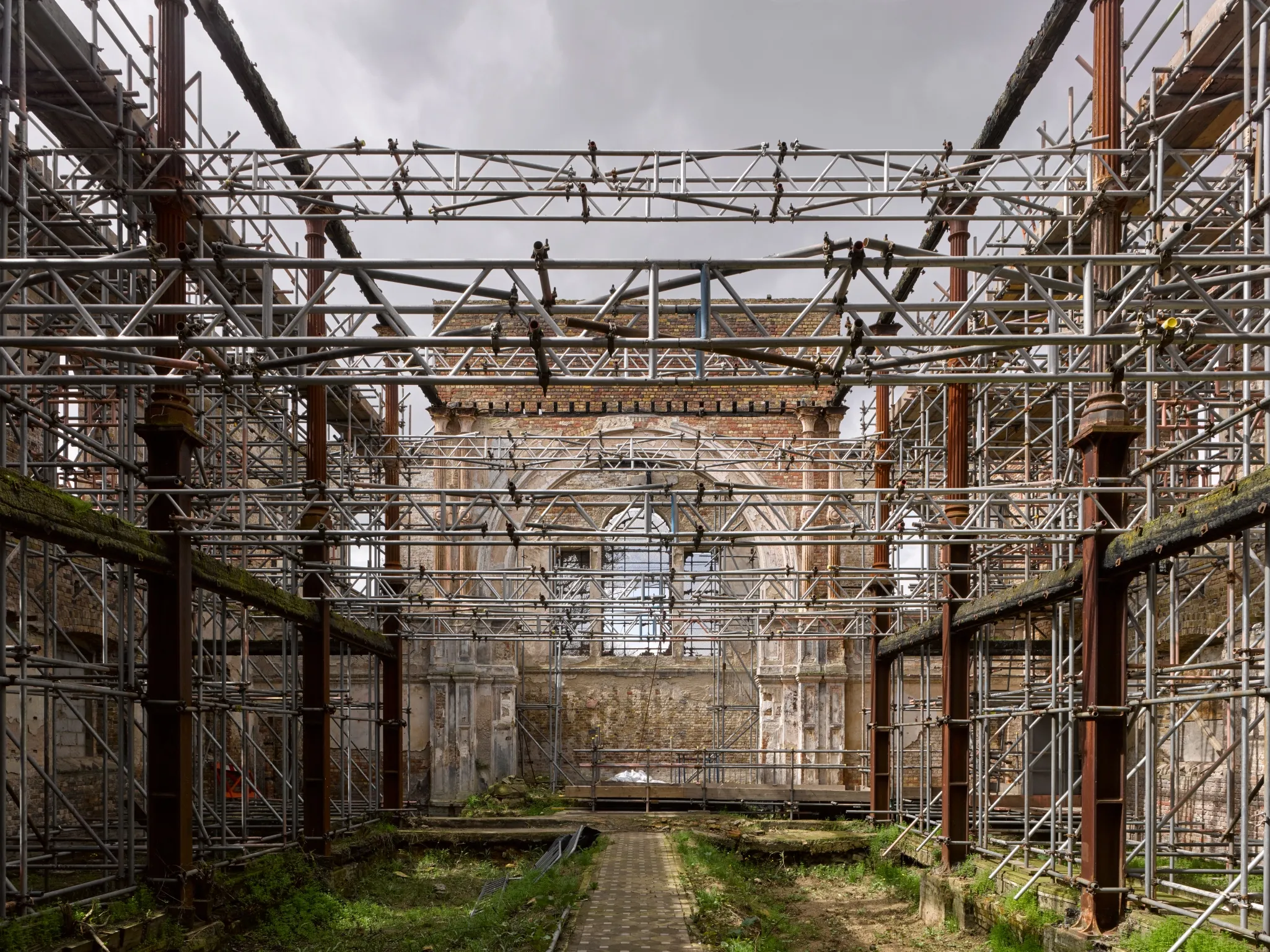 Interior of a large, partially restored building with exposed brick walls, metal scaffolding, and some overgrown vegetation.