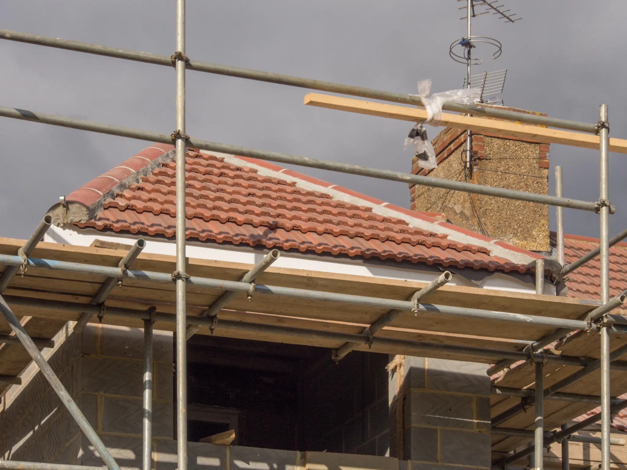 Scaffolding surrounds a house under construction with a red-tiled roof and a chimney. An antenna is mounted on the chimney.