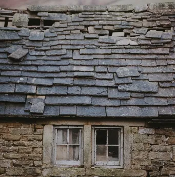 Old stone house with a deteriorating slate roof and two small windows, some shingles are missing or damaged.