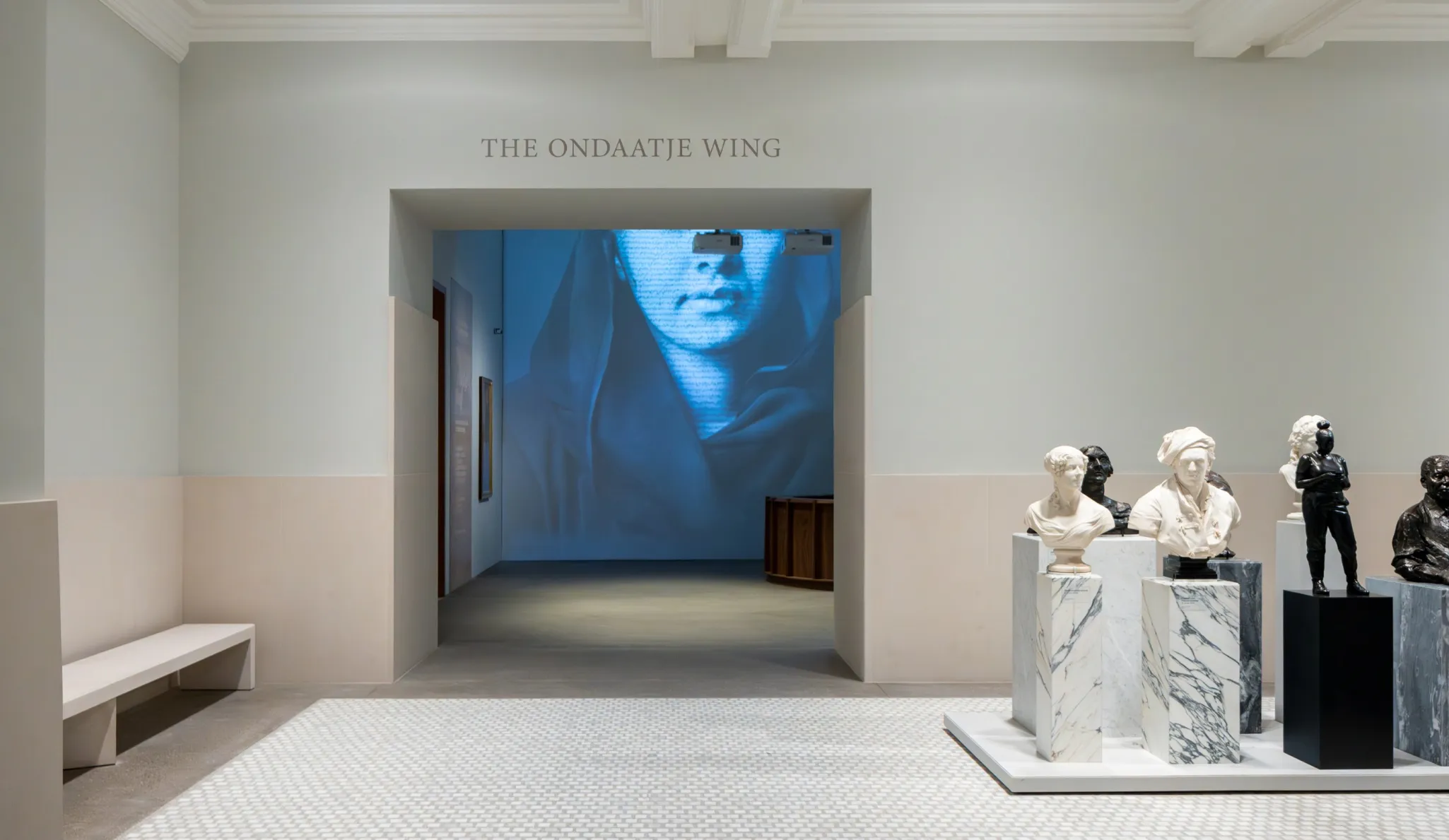 Museum gallery with sculptures and a marble floor. An adjacent room has a large blue-tinted digital projection of a woman's face.