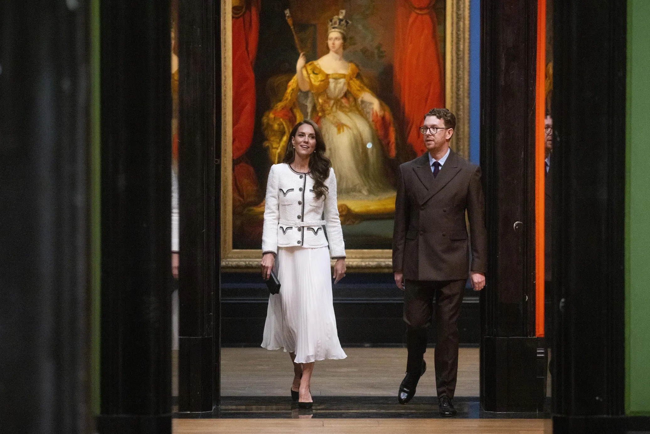 Two people walking in an art gallery with a large painting of a woman in a white dress and crown displayed behind them.