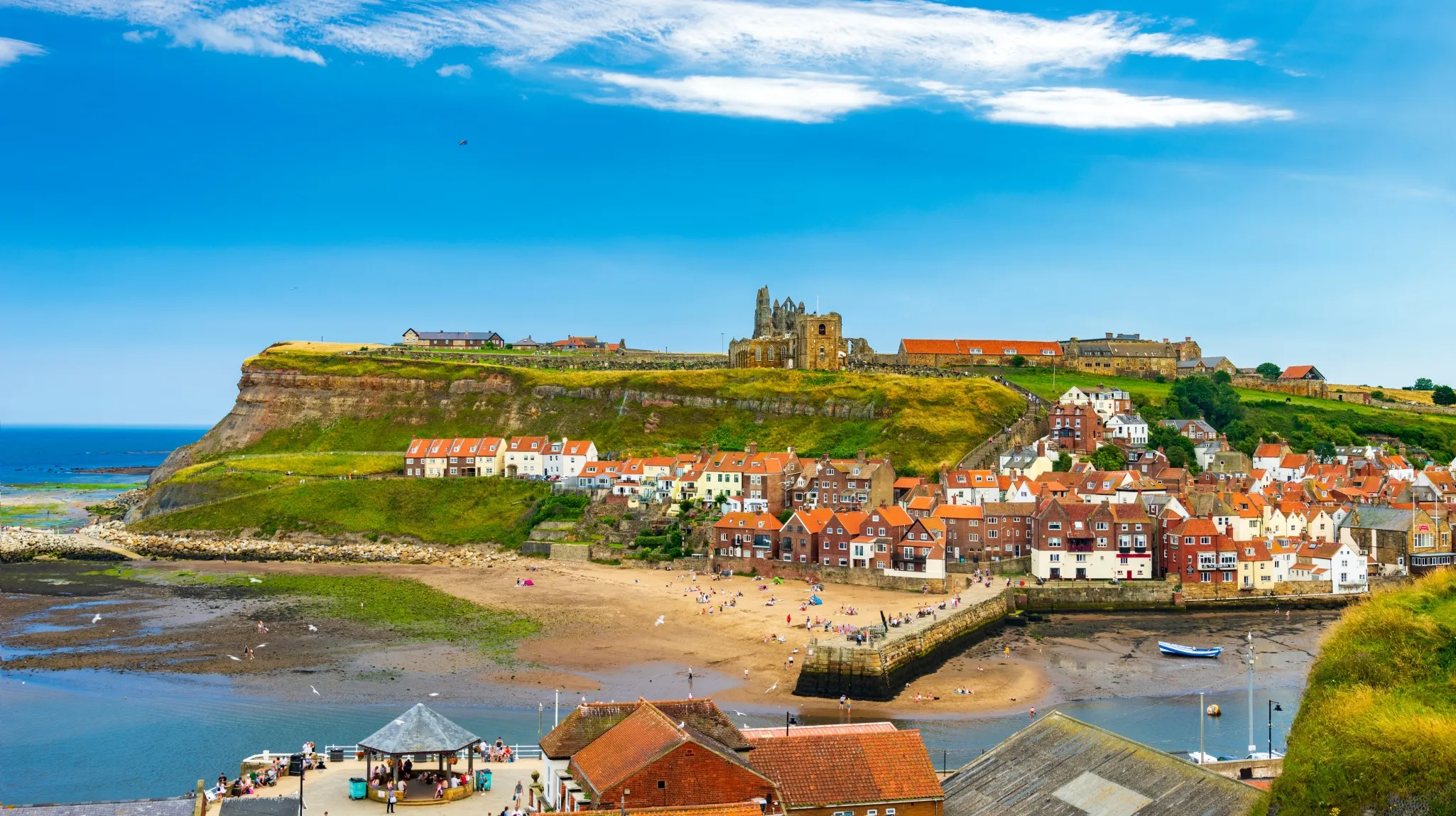 Colorful coastal town with a hilltop castle, quaint houses, and a sandy beach, under a vibrant blue sky with scattered clouds.