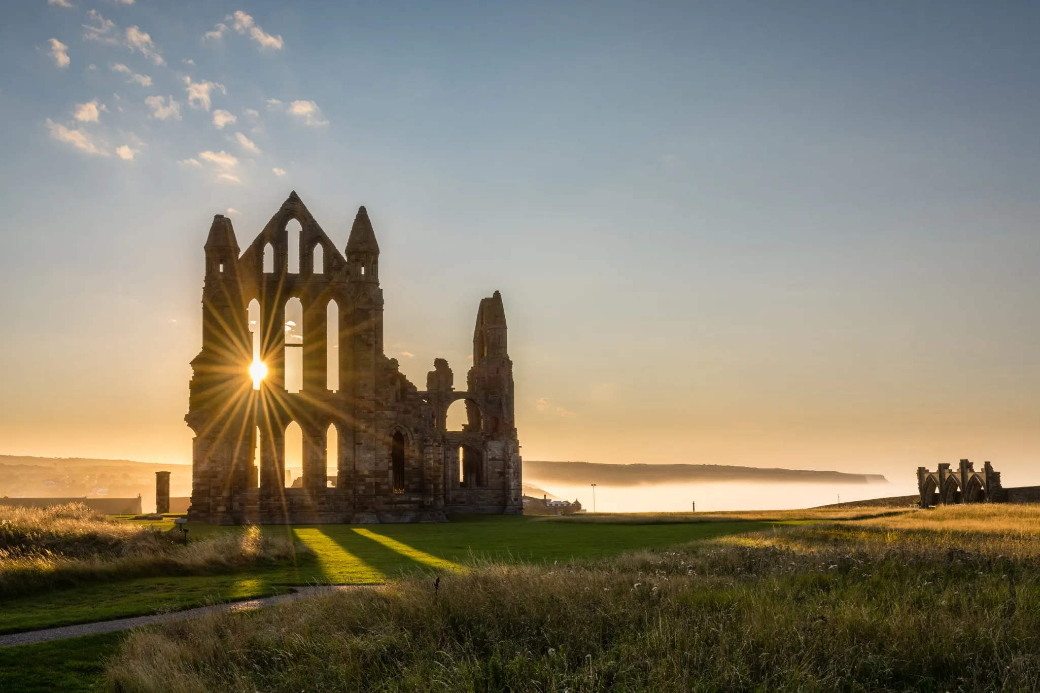 Sunrise illuminates the ruins of an ancient abbey, casting long shadows on the grassy landscape with a distant coastline in view.