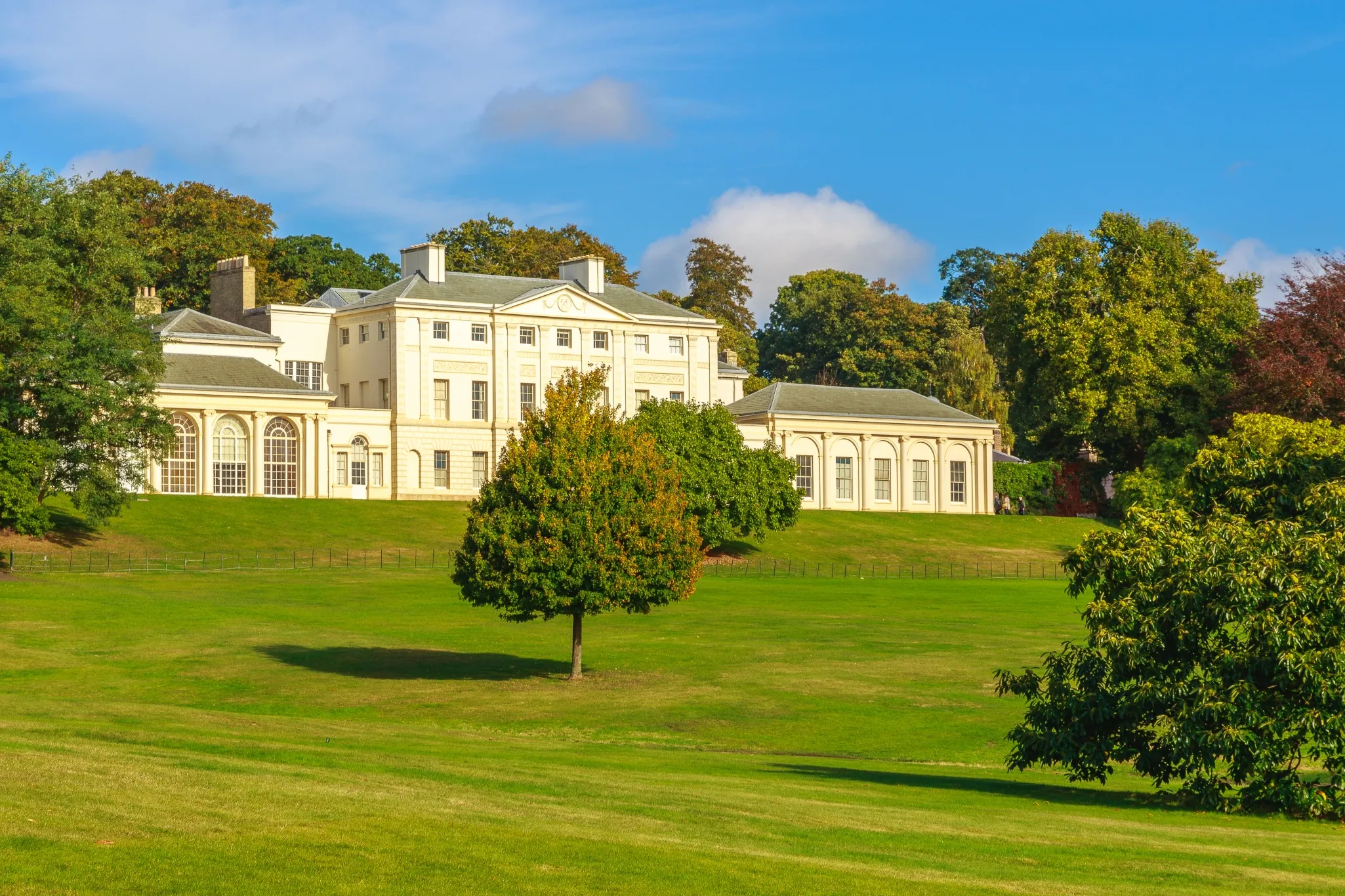 A large, cream-colored mansion surrounded by green lawns and trees under a clear blue sky.