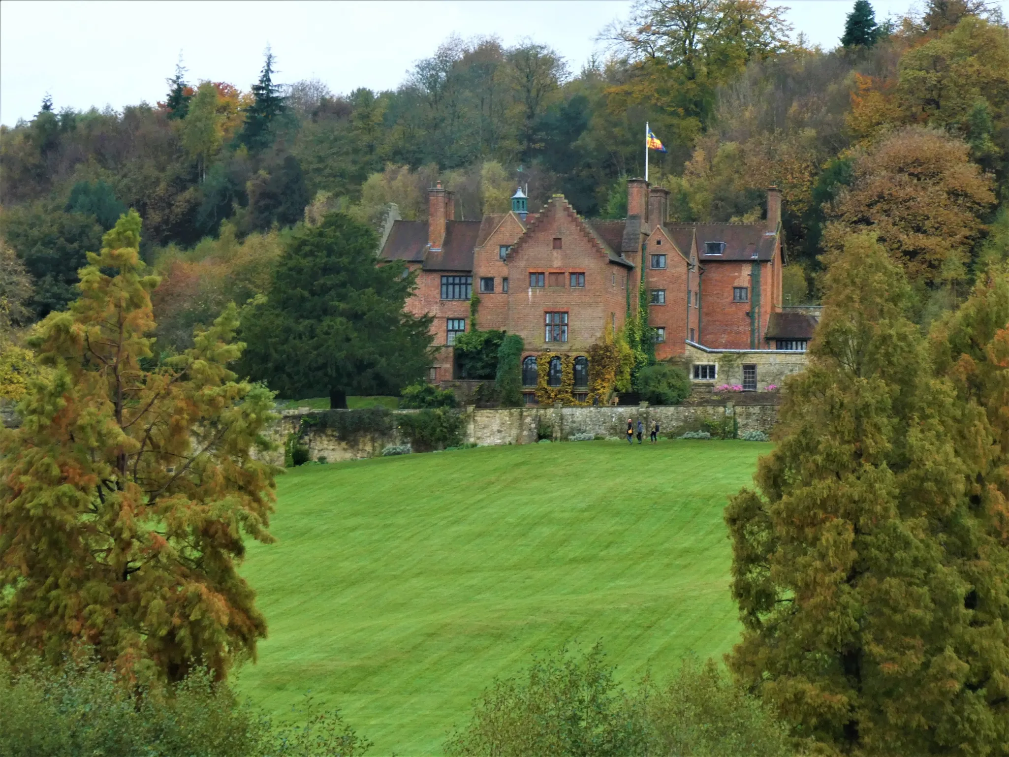 Historic brick mansion with a flag atop a turret, surrounded by a large green lawn and autumn trees, set against a forested backdrop.