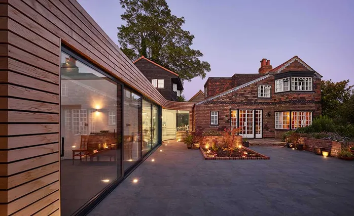 Modern wooden extension with large glass windows adjoining a traditional brick house, illuminated by outdoor lighting at dusk.