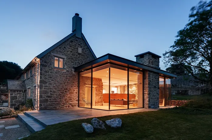 Modern stone house with large glass windows, illuminated from inside, stands amidst a natural setting during twilight.