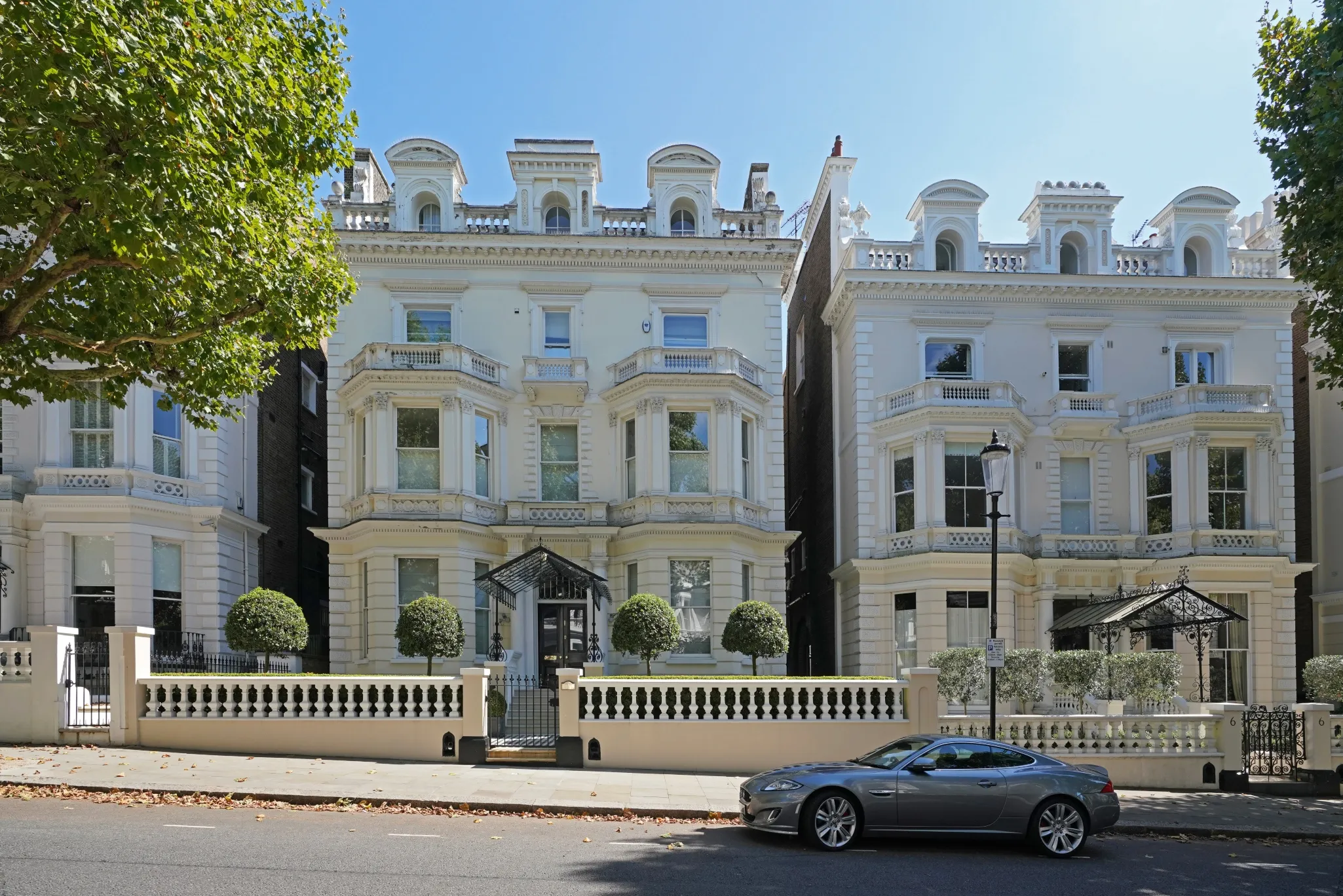 Exterior view of two ornate, multi-story buildings with a parked grey car and trees lining the street on a clear day.