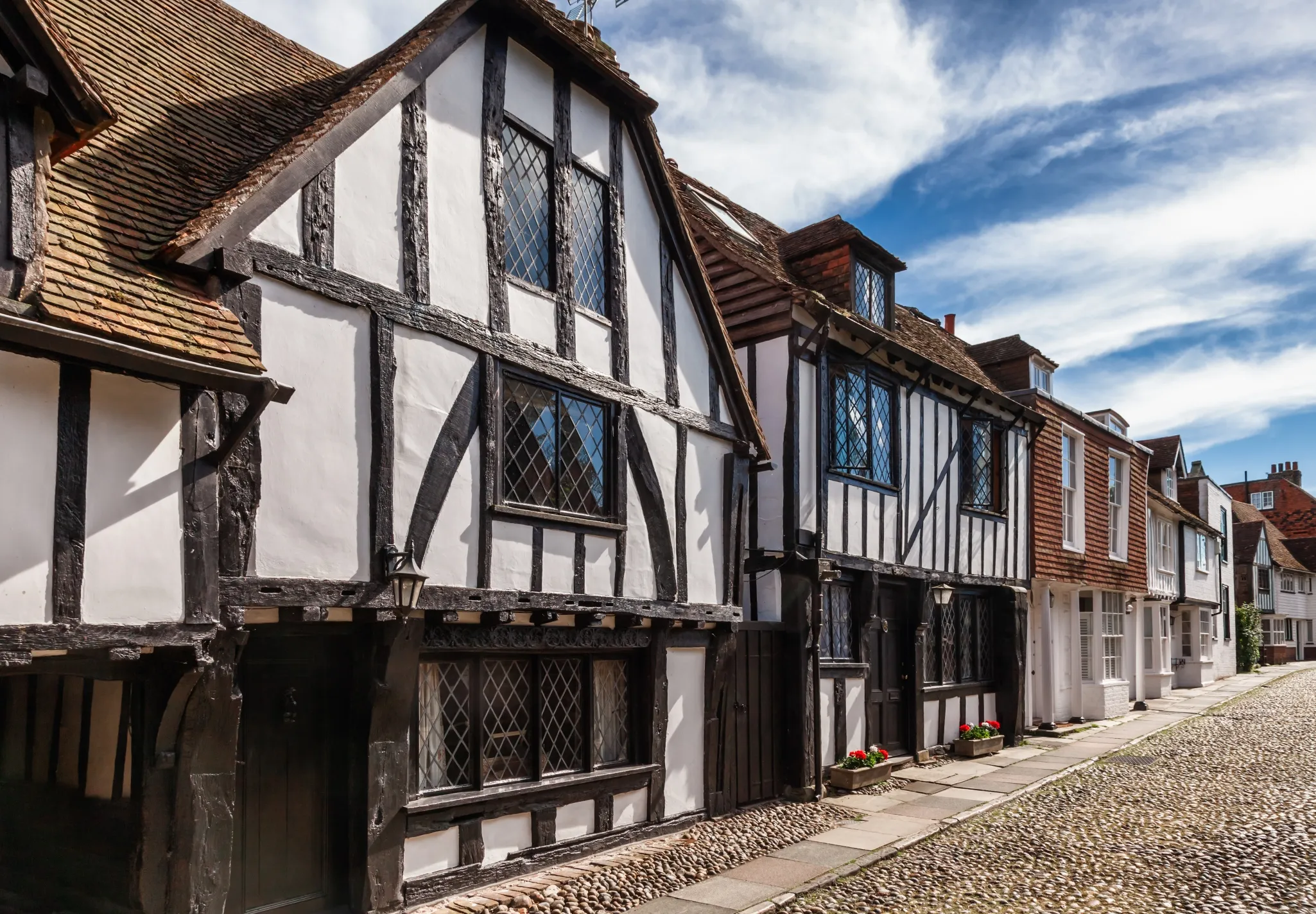 A row of historic half-timbered houses lines a narrow cobblestone street under a clear blue sky.