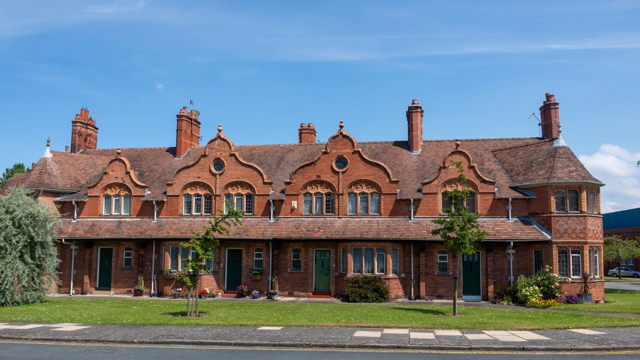 A red-brick row house with chimneys and decorative elements against a blue sky on a sunny day, featuring green doors and small trees in front.