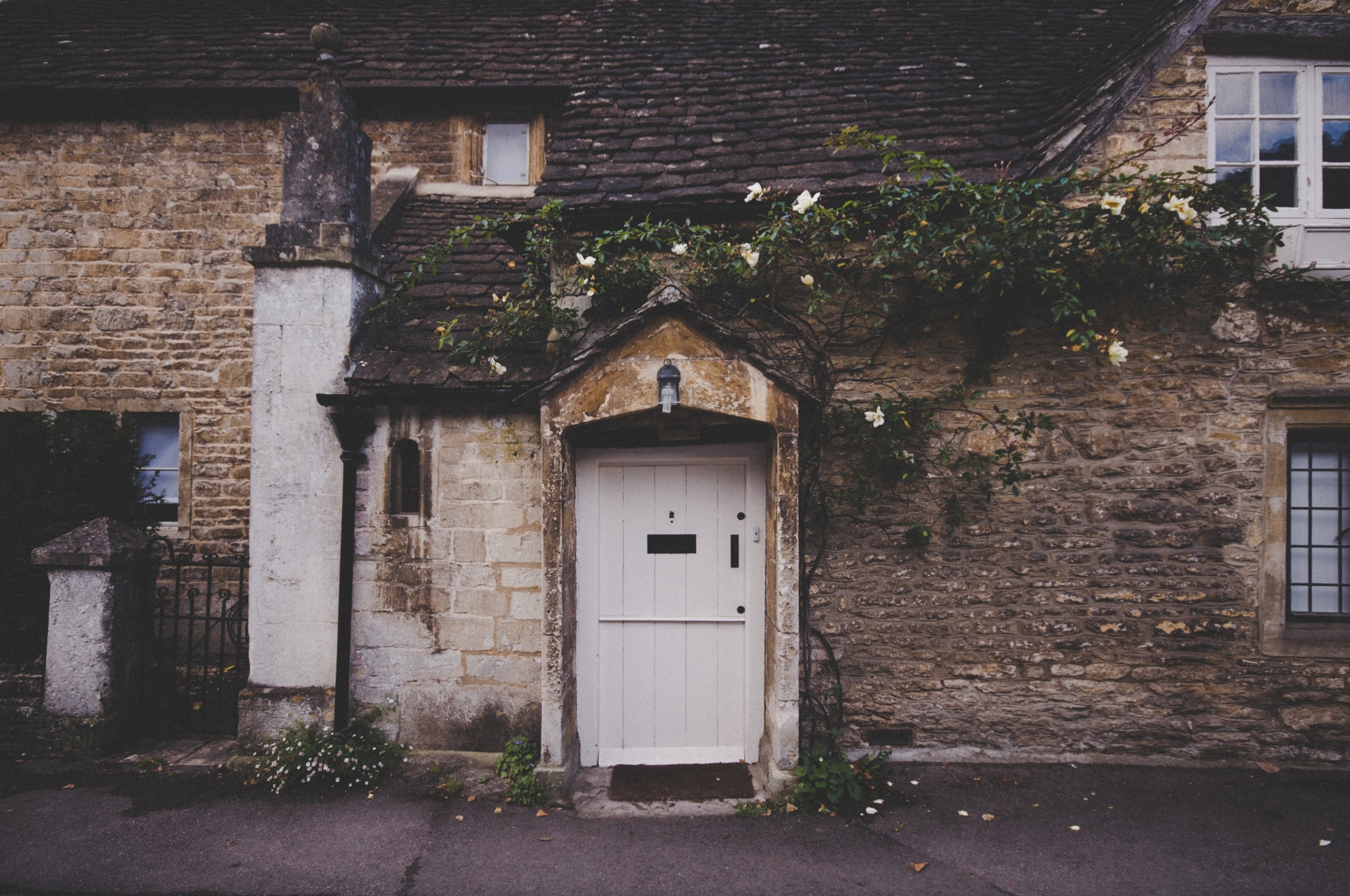 A quaint stone cottage with a white door, climbing plants, a small window, and a pitched roof, located on a quiet street.