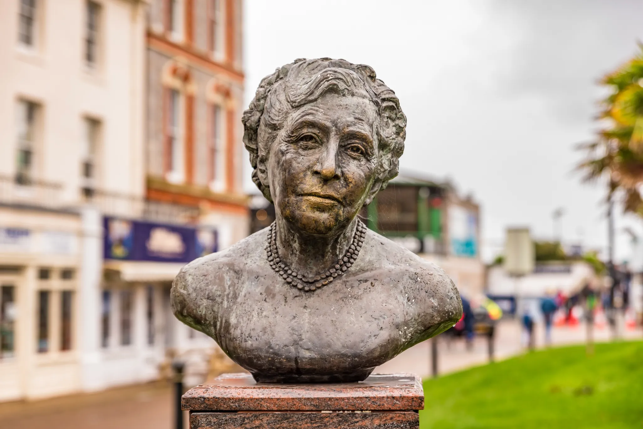 A stone bust of a woman with a pearl necklace is displayed on a pedestal in an urban outdoor setting with buildings and greenery.