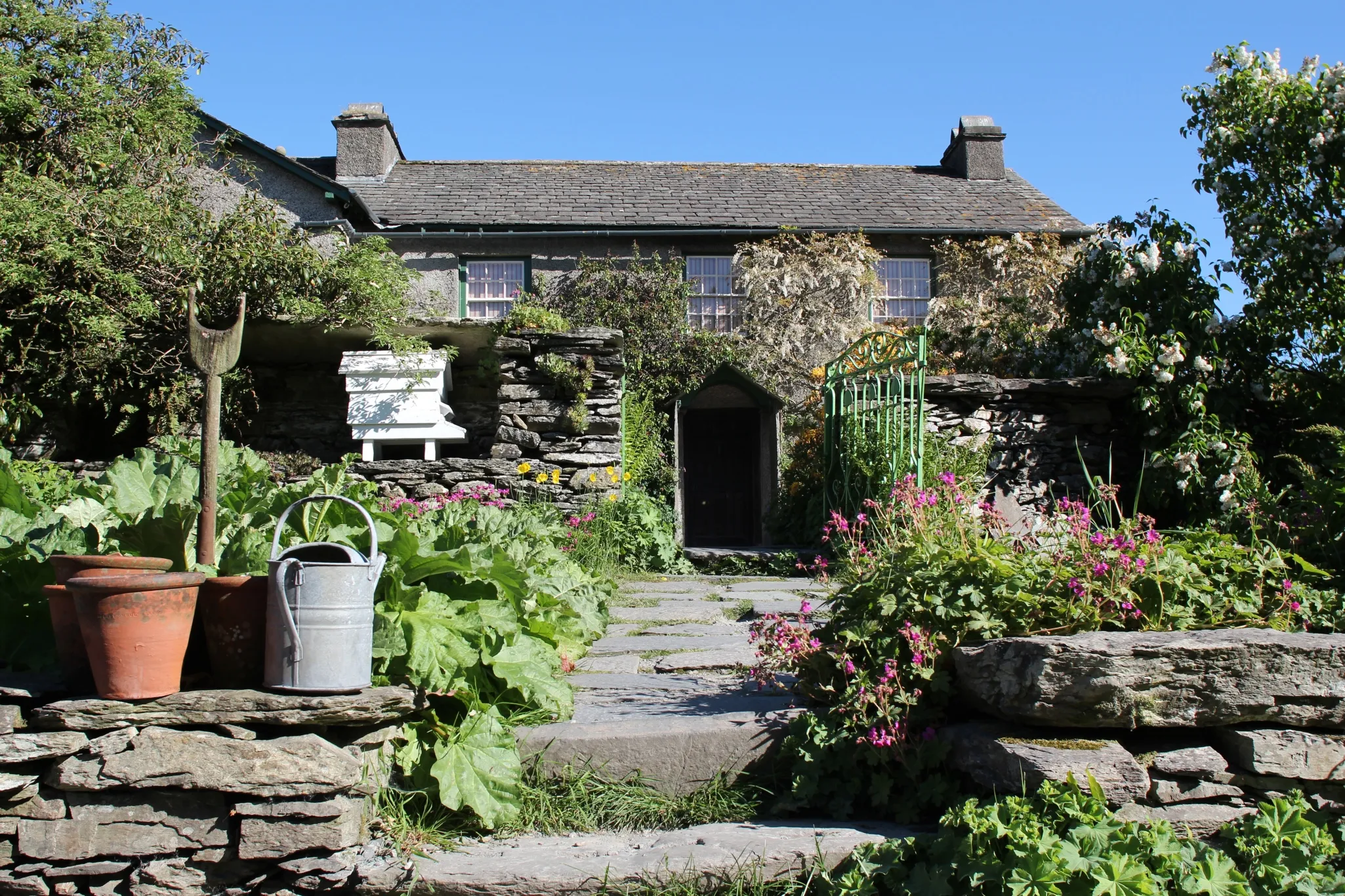 A stone pathway leads to a rustic house with a lush garden, featuring a watering can and a clay pot on the left side.