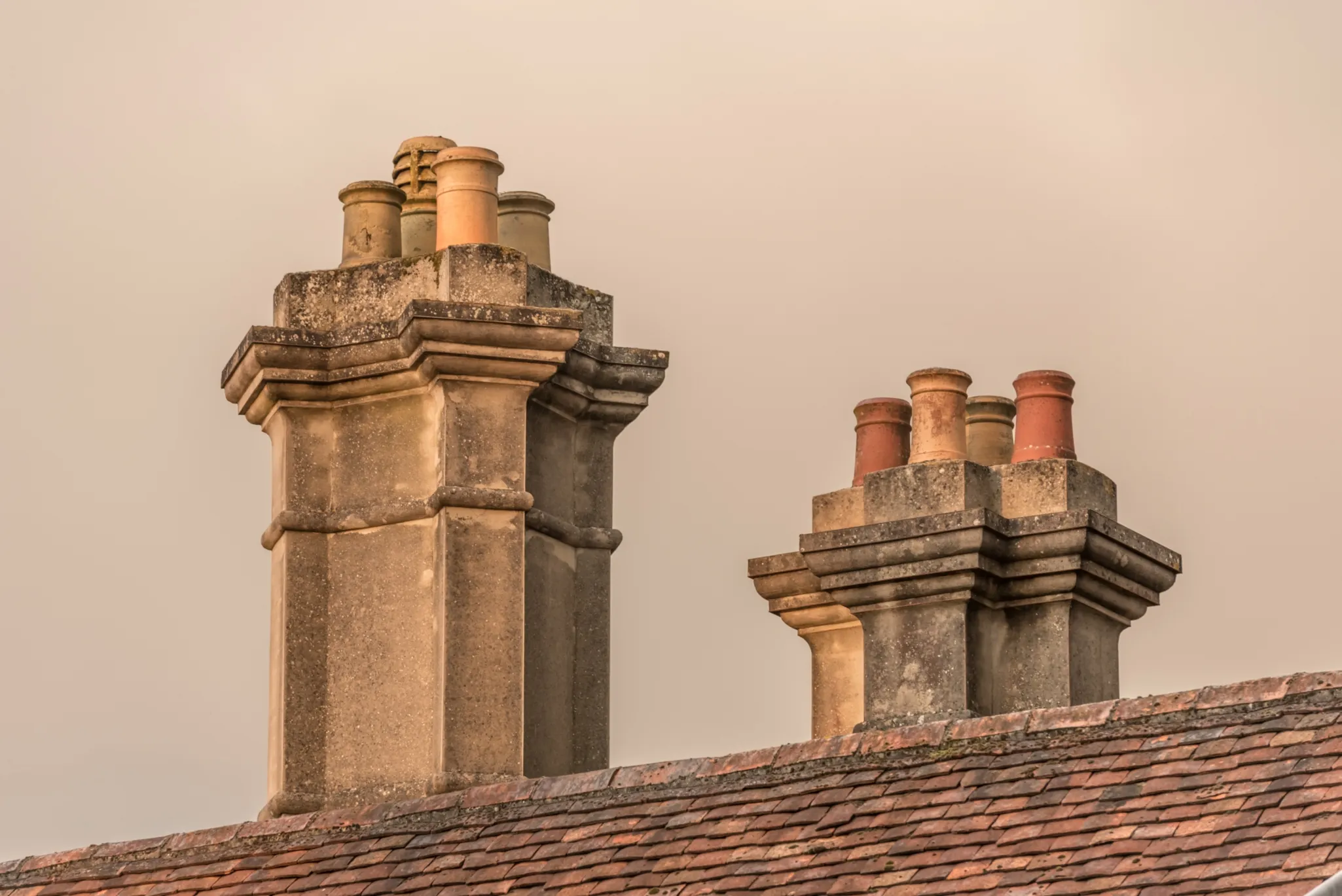 Two brick chimneys with multiple clay chimney pots on the roof of a building under a beige sky.