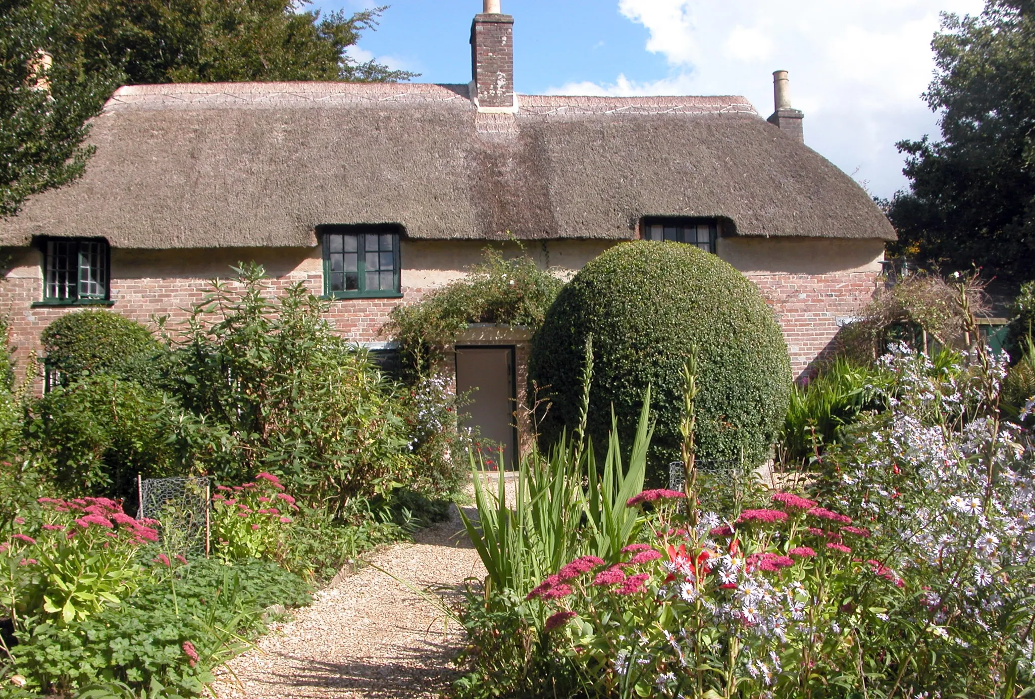 A quaint cottage with a thatched roof sits behind a lush, colorful garden with a gravel path leading to the front door.