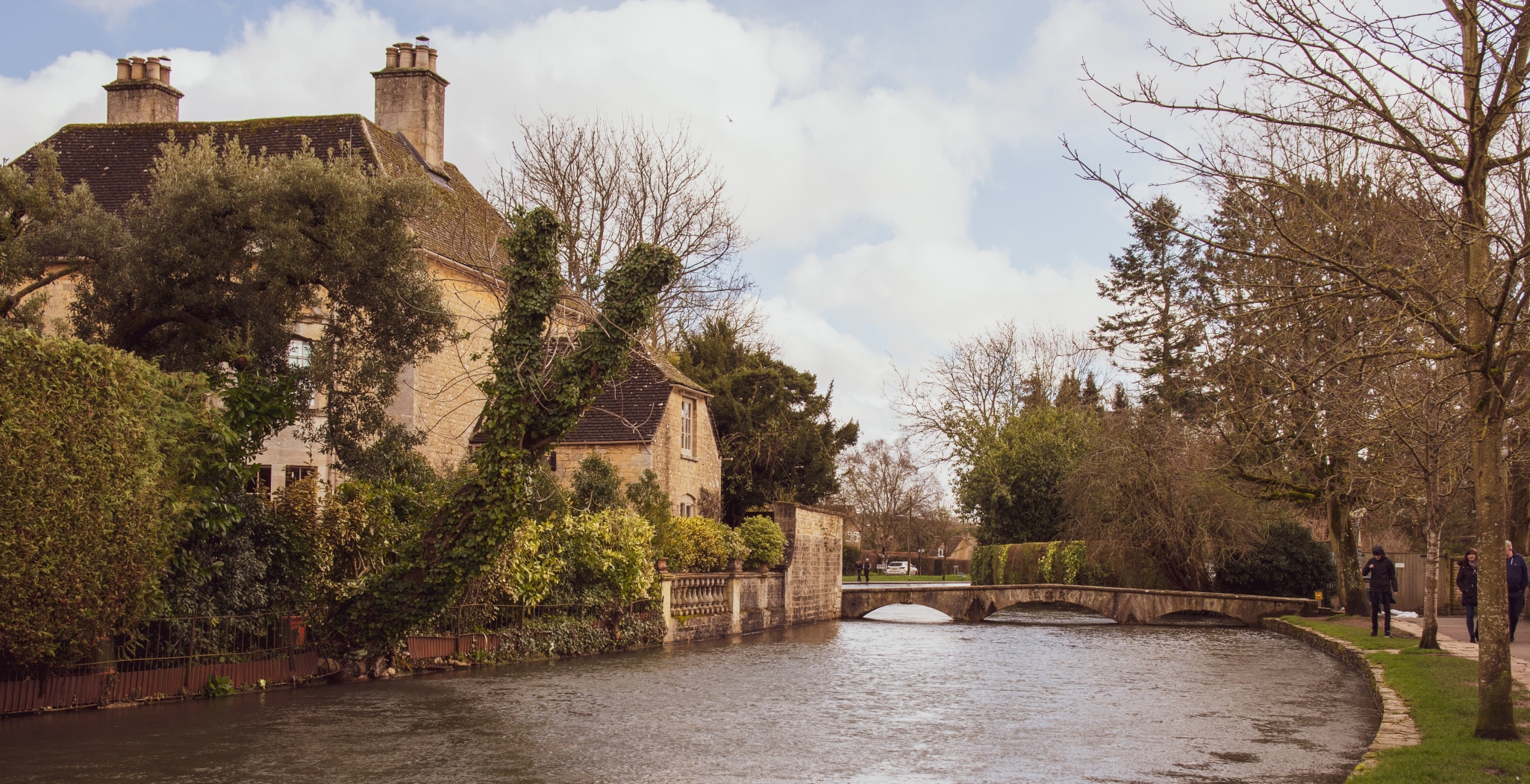 A riverside view featuring a stone house with lush foliage, a small arched bridge, and two people walking on a path beside the river.