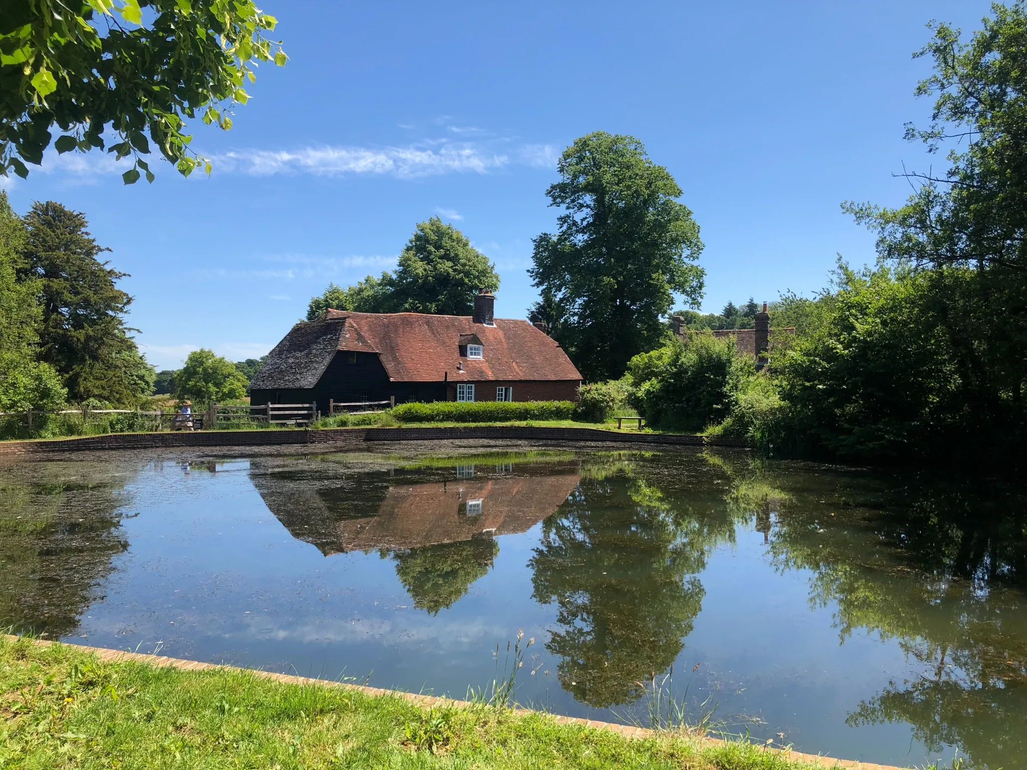 A quaint house with a red-tiled roof and a thatched extension is reflected in a clear pond surrounded by green trees and a blue sky.