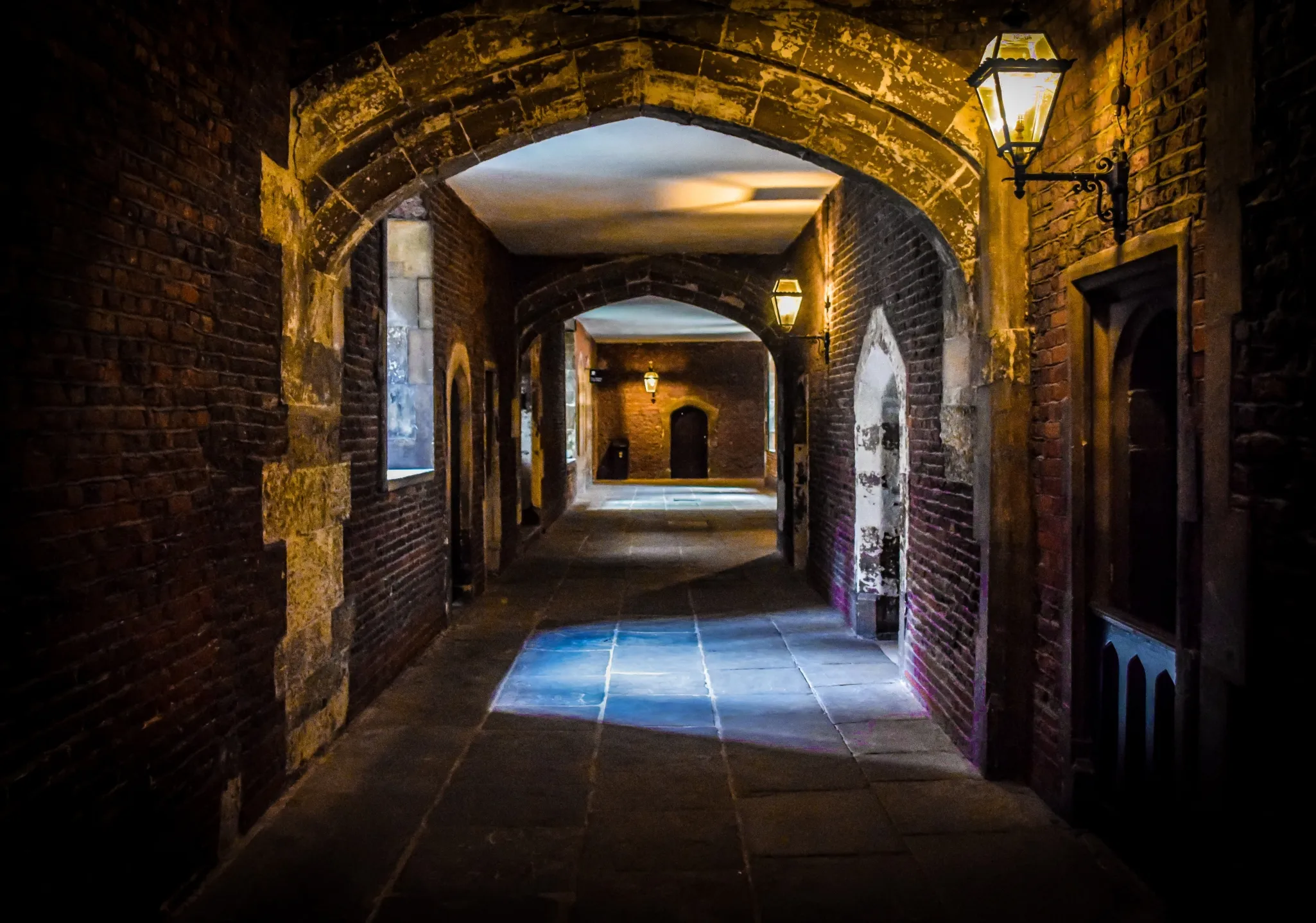 Dimly lit stone corridor with arched ceilings, illuminated by lanterns, and leading to a wooden door at the far end.